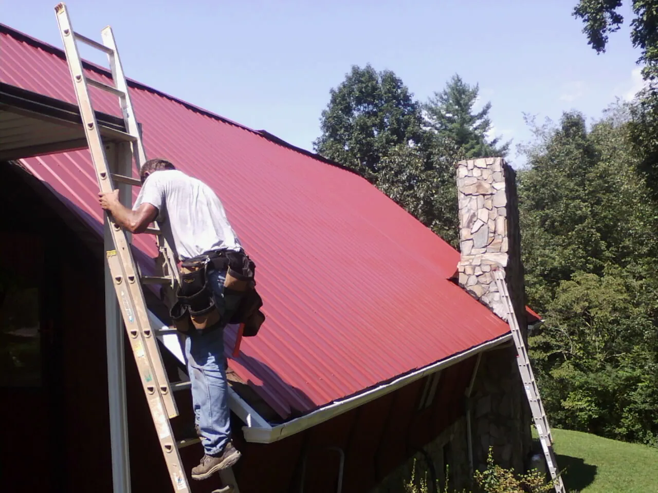 Roofer on a ladder beside a red metal roof and stone chimney — Soffit & Fascia Repair by Roofing Brothers in Pacific Grove